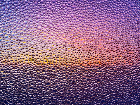 close-up of bead of condensation on a colored window in the background in shades of purple and yellow.