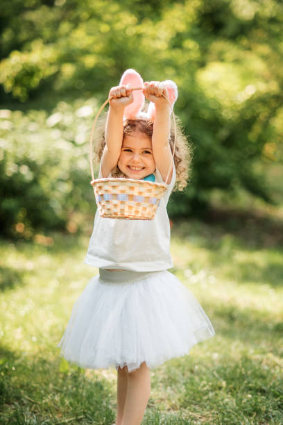 Easter egg hunt. Girl child Wearing Bunny Ears Running To Pick Up Egg In Garden. Easter tradition. Baby with basket full of colorful eggs.