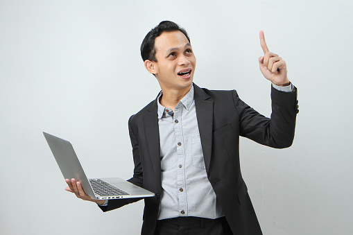 Happy asian indonesian business man in suit holding laptop computer with pointing finger gesture on isolated background