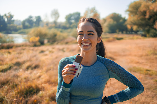 A satisfied young female athlete having a protein bar for a snack on her break from a routine jog in nature.