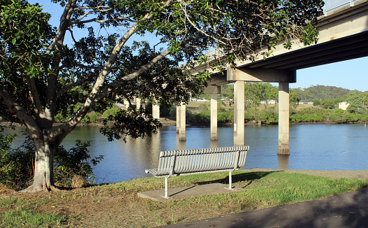View of the John Oxley Bridge, Boyne River, a tree and a metal park bench seat at Tannum Sands, Queensland, Australia
