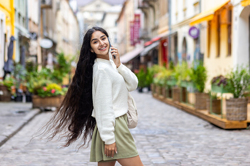 Portrait of a beautiful young Indian woman with long hair walking on a city street, talking on the phone, posing and looking at the camera.