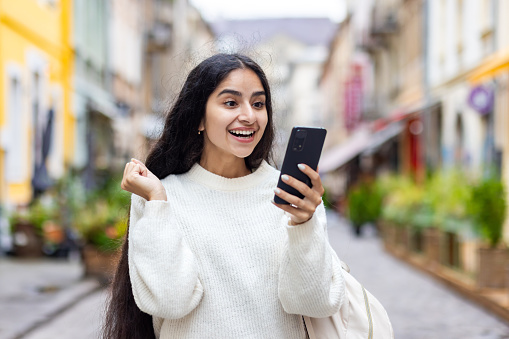 Close-up photo of happy Indian young woman standing on city street and looking at phone screen, happy to receive news and message showing victory gesture with hand.