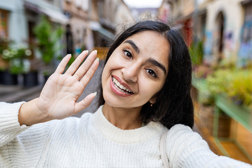 Happy young Indian woman tourist waving hello during a city walk. She enjoys sightseeing and exploring new places with a smile.