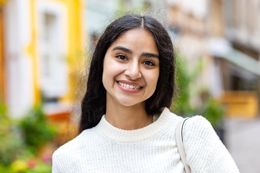 Close-up portrait of a beautiful and smiling Indian woman standing on the street in a white sweater and looking at the camera.