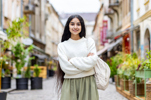 Portrait of a confident Indian young female student in a white sweater standing in the middle of a city street with her arms crossed on her chest and looking at the camera with a smile.