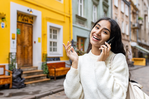 Close-up portrait of a beautiful Indian young woman, tourist and student talking on the phone emotionally, standing on a city street and looking at the camera.