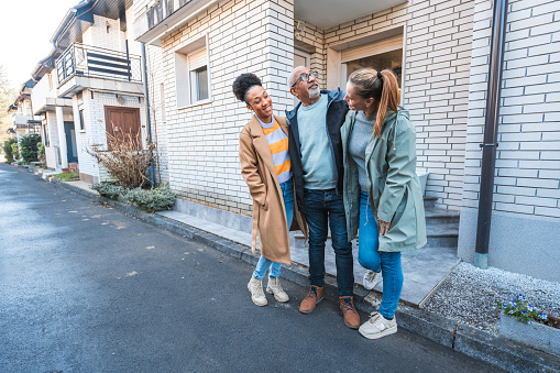 A joyful mixed race family of four stands close together, smiling outside their residential home. Two female and two male family members, dressed in casual attire, express happiness in an urban setting. They are engaged in a friendly conversation, enjoying a sunny day.