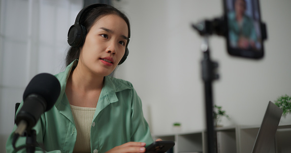Selective focus, Happy young woman wears headphones talk speech into microphone share information record for internet audience video blog on smartphone in home studio. podcast concept
