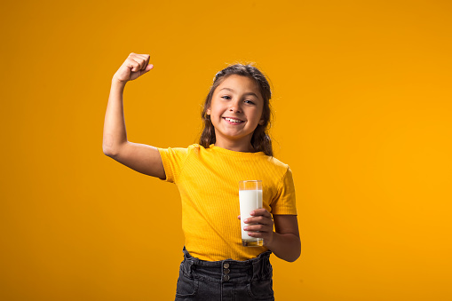 Portrait of smiling child girl holding glass of milk and showing strenght gesture. Nutrition and health concept