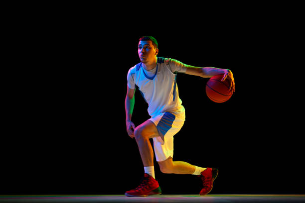 istock Skilled, focused basketball player performs combined ball-handling technique against black studio background in mixed neon light. 2029873016