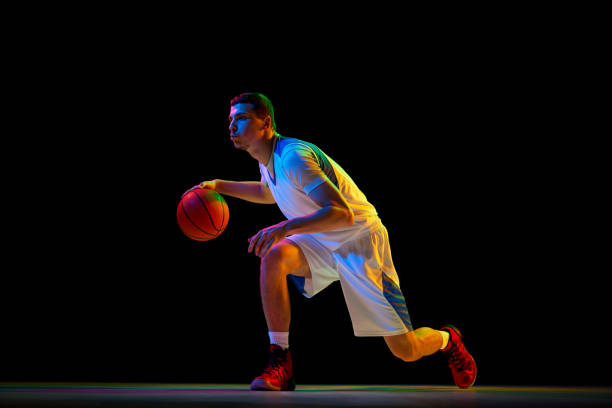 istock Young, focused basketball player performs low-handling technique with covering ball against black studio background in mixed neon light. 2029873167