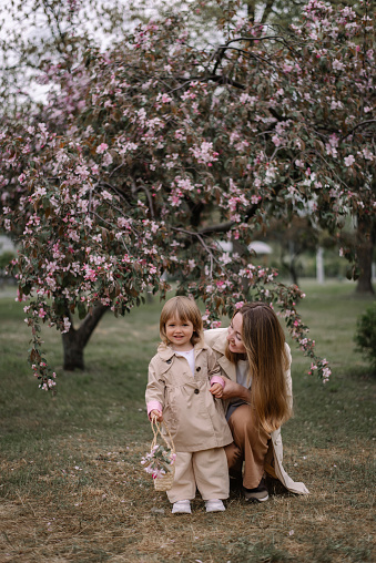 A little girl with her mother against the background of an apple tree blooming with red flowers. Girl and mother in stylish light clothes. The girl has a basket of apple flowers in her hands