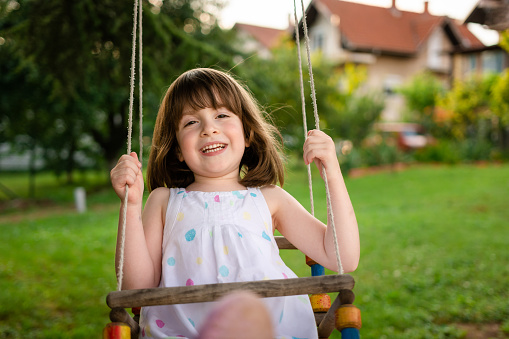 A happy little girl is swinging on a swing in the yard and looking at camera