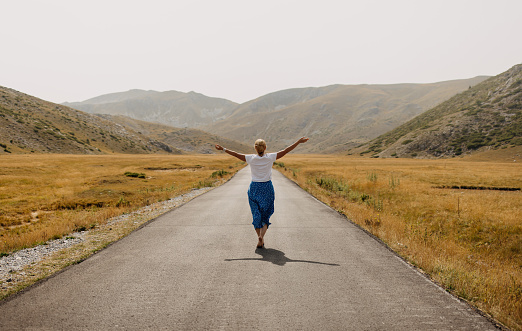 One woman wearing blue skirt and white t shirt walking barefoot on empty mountain road with arms outstretched