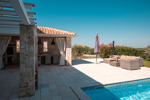 Wide angle shot of a Mediterranean-style villa with a patio and a pool. Sun beds and  shades ready for use, a set of  patio furniture in one corner. Strong summer light and contrasting shadow from the house.