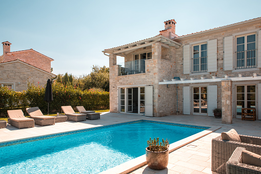 Wide angle shot of a stone villa in Mediterranean countryside. A large pool and sunbeds for multiple guests. Potted bush and wicker lounge chairs on the terrace in front of the house.