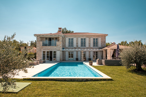 Wide angle shot of a 5-bedroom holiday house in countryside, large lawn and a swimming pool attached to the house. Blue sky and summer light.