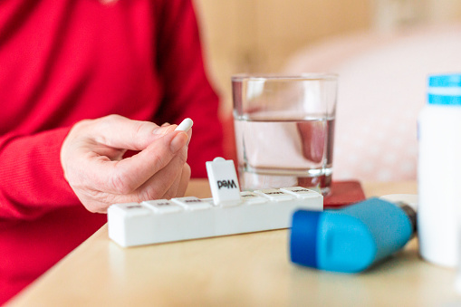A senior woman sitting on the edge of her bed at home, about to take her medication from the pill box on her bedside table. There is also an asthma inhaler on the table.