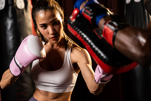Asian female boxer punching in fierce boxing training session, delivering strike to her sparring trainer wearing punching mitts, showcasing boxing technique and skill. Impetus
