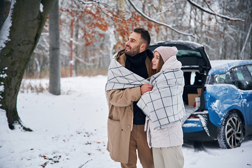 With warm towel. Happy couple having a walk in winter forest. Blue car is parked.
