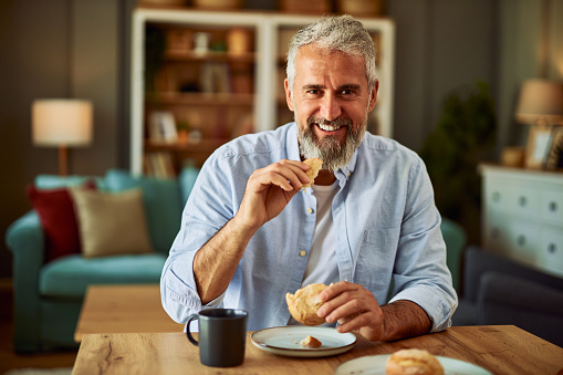 A portrait of a smiling senior adult man with beard eating a delicious pastry at the dining table in a cozy apartment on a lovely, sunny morning.