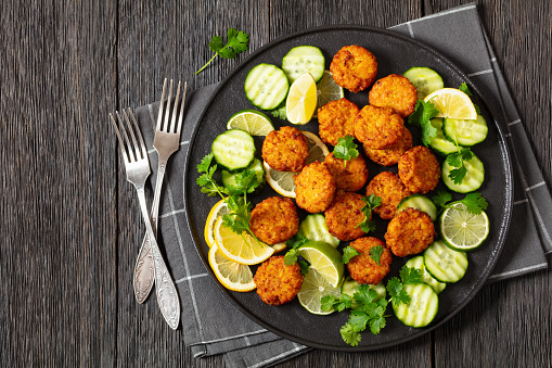 fried rice cutlets, rice tikki, indian style on black plate served with cucumber, lemon, lime slices and forks on dark wooden table, flat lay, free space
