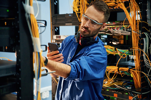 Holding smartphone. Young man is working with internet equipment and wires in server room.