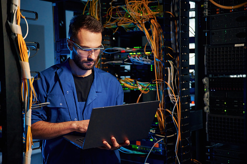 In glasses. Young man is working with internet equipment and wires in server room.