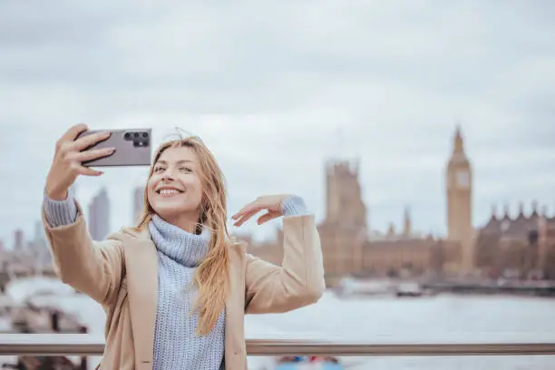 Candid waist up horizontal portrait of a young cheerful good looking blond woman in her 30s taking selfie on her smart mobile phone device while laughing with toothy smile during visiting and exploring down town capital city of London, United Kingdom. Selective focus with priority on the model with plenty of copy space on the background and sky, which is defocused Westminster Bridge, Parliament building and Big Ben clock tower. Photo created during cold season outdoors and the model is with warm casual clothes in light cream and blue colours on a cloudy day  - creative stock photo