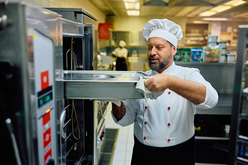 Mature chef using professional oven while preparing food in a restaurant.