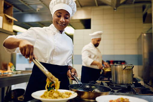 Happy African American professional cook serving a dish while working in a restaurant.