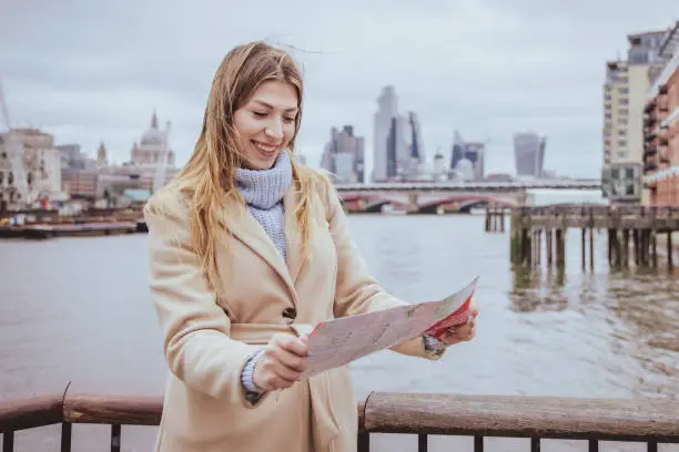 Candid wide angle portrait of a young European tourist woman in her 30s with toothy smile looking down, while holding a printed city map while exploring the capital city of London, United Kingdom. Selective focus on the model with defocused skyline at the back including the iconic Saint Paul cathedral and corporate finance business buildings at Liverpool street, photo is horizontal with plenty of copy space in the sky and background. Shot on full frames camera during a cold season with casual warm clothes in light cream and blue. Colours - creative stock image