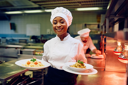 Happy African American female chef serving a dish in restaurant and looking at camera.