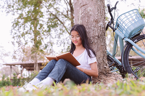12 year old Asian girl wearing eyeglasses and a white shirt sitting reading a book under a tree in the park with blue bicycle, Young woman with Mental health and relaxation in nature concept.
