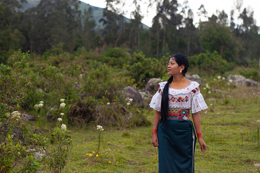 A woman adorned in a traditional embroidered blouse and skirt strolls thoughtfully across a grassy field, with a backdrop of mountains under a softening sky suggesting dusk. She appears serene, connected with the natural surroundings of this rural setting.