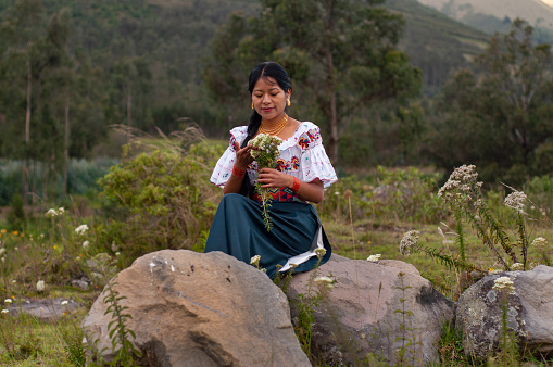 A woman dressed in traditional indigenous attire sits on a large stone in a natural setting, holding a bouquet of freshly picked wildflowers. She is caught in a moment of serene contemplation, reflecting the essence of Earth Days connection with nature and cultural heritage.