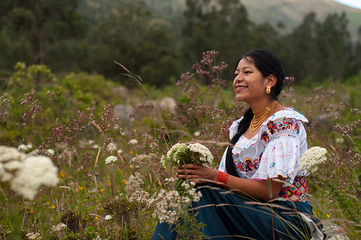 A smiling indigenous woman, wearing traditional embroidered clothing and jewelry, sits amidst wildflowers in a mountain meadow. She holds a bunch of freshly picked flowers and appears to be enjoying a peaceful moment in nature as dusk settles over the landscape.