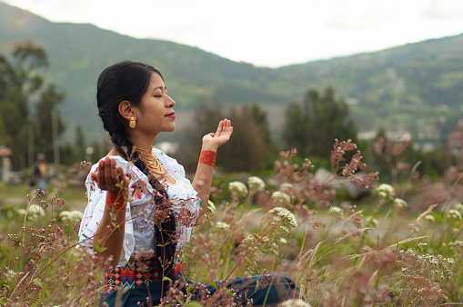 A woman dressed in vibrant traditional clothing sits cross-legged in a field of wildflowers, her eyes closed in meditation. The soft light of the setting sun enhances the tranquil atmosphere of the rural landscape.