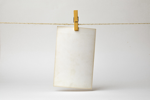 An empty, aged photo paper note is suspended from a twine using wooden clothespins against a neutral, seamless backdrop, suggesting a message or reminder.