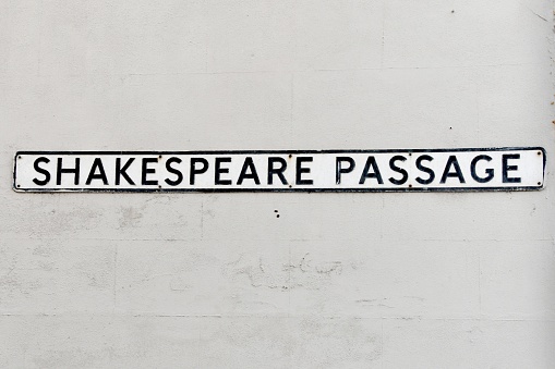 close up of Shakespeare Passage street  sign.  The plaque is black text on white metal against a grey concrete wall background outside