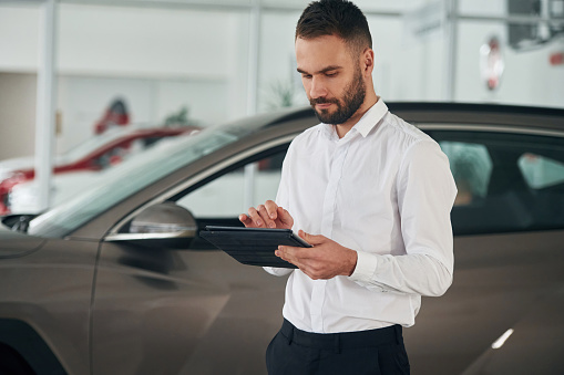 Black colored tablet in hands. Worker in formal clothes is in the auto salon.