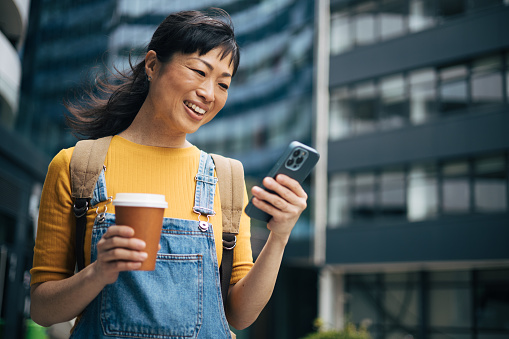 Yapanese woman holding a smartphone and a cup of coffee looking at device screen against the background of an office building