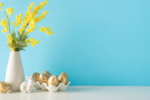 istock Kitchen adorned for Easter, side view shot, with a table bearing a ceramic holder filled with golden eggs, a hare figurine, next to a mimosa vase, against a faint blue wall 2030246624