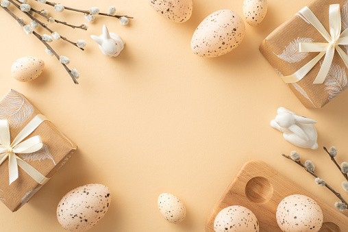 Quaint Easter setup visualization. Top view of beige paper presents, bunny figures, dotted quail eggs in wooden holder, pussy willow, all positioned on soft beige backdrop with space for text or ads