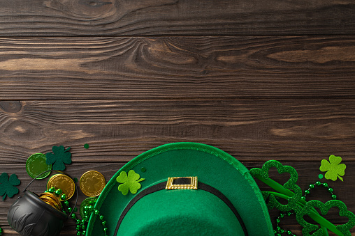 Spellbinding St. Patrick's scenario from top view, filled with shamrocks, leprechaun headwear, currency coins, treasures, party lenses, garlands, positioned on wooden tableau, space for text or promos