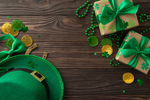 Festive St. Patrick's theme arrangement overhead, highlighting shamrocks, gnome hat, riches coins, presents, bowtie, lucky horseshoe, chains, glitter, laid out on wooden surface, space for promotions