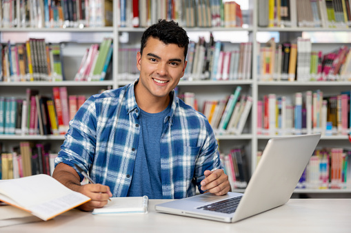 Happy Latin American man studying at the library and looking at the camera smiling - education concepts