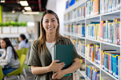 Portrait of a happy post-graduate student smiling at the library holding a book and looking at the camera