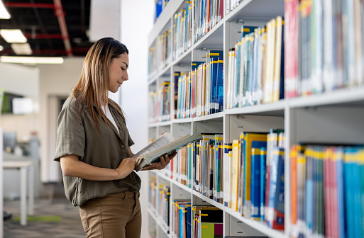 Happy Latin American woman searching for a book at the library - education concepts